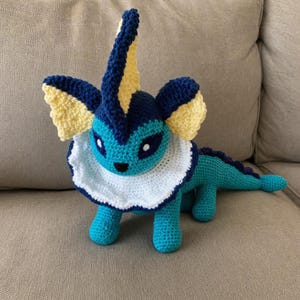 May include: A handmade crochet plush toy shaped like a creature with a teal body, a white ruffled collar, and dark blue and pale yellow accents on its head and ears. The figure has a pointed crest, large dark eyes, and a long tail with a fin.