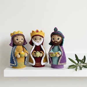 May include: Three crocheted figures representing the Three Wise Men, each wearing a crown and a cape. The figures are dressed in yellow, maroon, and light blue garments with purple and green accents, holding small containers with beads and gems.