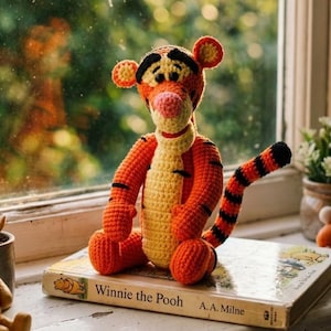 May include: A crocheted Tigger plush toy with orange and yellow yarn, black stripes, and a pink nose, sitting on a hardcover book titled Winnie the Pooh by A.A. Milne. The stuffed animal features round ears, a long striped tail, and stitched eyes.