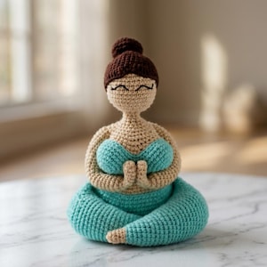 Puede incluir: Una muñeca de ganchillo con piel bronceada, cabello castaño oscuro recogido en un moño y una cara sonriente con los ojos cerrados. La muñeca viste un top turquesa y pantalones turquesas, sentada en posición de loto con las manos juntas.