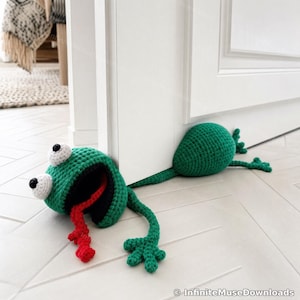May include: A green crocheted frog doorstop featuring a large head with white eyes, black pupils, and a long red tongue. The body is flattened and extends underneath a white door, with green limbs and webbed feet visible on the floor surface.