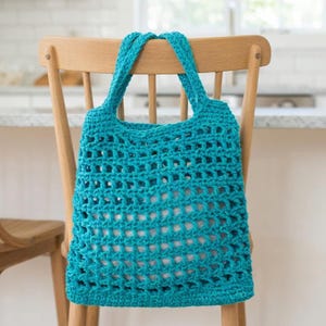 Puede incluir: Un bolso tote turquesa tejido a ganchillo, de forma rectangular y con dos asas. El bolso tiene un tejido abierto, similar a una red, y está colgado en el respaldo de una silla de madera. El bolso está vacío y parece hecho a mano.