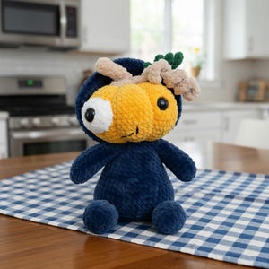 May include: A handmade plush toy in the shape of a whimsical creature. It features a navy blue body, a yellow head with a large white and black eye, and tan and green accents. The toy is sitting on a blue and white checkered tablecloth.