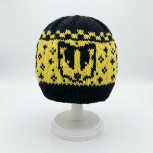 Könnte beinhalten: Eine Strickmütze mit schwarzem Rippbündchen und einer gelben Krone mit schwarzem geometrischem Muster. In der Mitte der Krone befindet sich ein großer, stilisierter schwarzer Buchstabe W, umgeben von kleineren rautenförmigen Motiven.