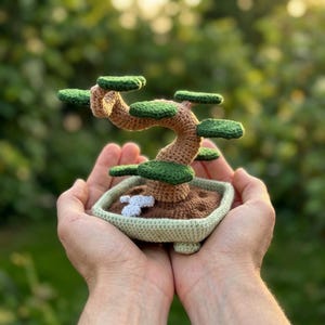 May include: A crocheted bonsai tree with a tan trunk and dark green foliage, planted in a light green square pot. The base of the tree features brown soil texture and small white stones, all crafted with yarn in a detailed, handmade style.