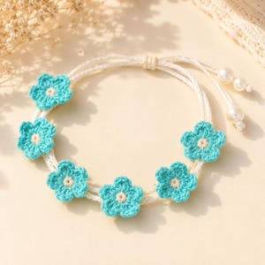 Può includere: Un braccialetto fatto a mano con sei fiori all'uncinetto turchesi con centri color crema. I fiori sono infilati su un cordoncino bianco, con una chiusura a nodo scorrevole e perle di perle. Questo braccialetto floreale è un accessorio affascinante.