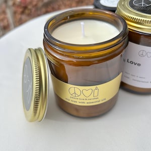 May include: A brown glass jar candle with a gold lid and a white wick. The candle has a yellow label with the text "peace love & candles" and a peace sign, heart, and candle icon. The lid is off to the side.