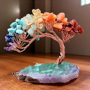 Könnte beinhalten: Eine dekorative Baumskulptur mit einem Kupferdrahtstamm und Ästen, verziert mit farbenfrohen Edelsteinen. Die Edelsteine sind in einem Regenbogenspektrum angeordnet, und die Basis ist eine Mischung aus grünen und violetten Kristallen. Die Skulptur steht auf einer Holzoberfläche.