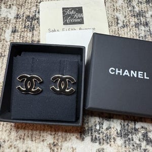 Op de afbeelding: Goudkleurige oorbellen in een zwarte doos met het CHANEL-logo. De oorbellen hebben de vorm van in elkaar grijpende C's. Een kassabon van Saks Fifth Avenue is zichtbaar op de achtergrond.
