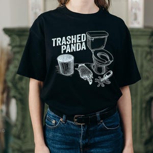 May include: Black t-shirt with a white graphic design featuring a bin, a raccoon, a toilet, and a spilled bottle. The text "Trashed Panda" is above the graphic. The shirt is paired with blue jeans.