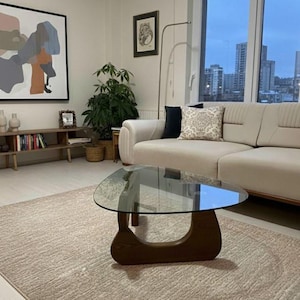 Könnte beinhalten: Ein modernes Wohnzimmer mit einem hellen Sofa mit dekorativen Kissen, einem einzigartigen Couchtisch aus Holz mit Glasplatte und einem großen abstrakten Gemälde. Der Raum hat eine neutrale Farbpalette und natürliches Licht.