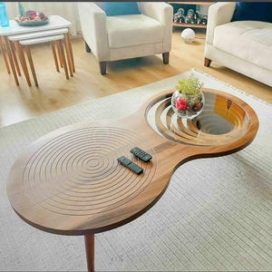 Puede incluir: Una mesa de centro de madera con una tapa de cristal transparente y un diseño circular. La mesa tiene un acabado marrón oscuro y está sostenida por tres patas de madera. Dos mandos a distancia negros están sobre la mesa. Un cuenco de cristal con flores está sobre la tapa de cristal.
