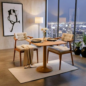 May include: A modern dining set featuring a square wooden table and two chairs. The chairs have patterned cushions and wooden frames. A beige throw is draped over one chair. The set is on a light-colored rug, with a city view in the background.