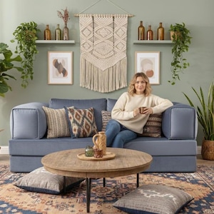 Puede incluir: Un sal&oacute;n acogedor con un sof&aacute; azul, cojines decorativos, una mesa de centro redonda de madera y una alfombra estampada. La habitaci&oacute;n est&aacute; decorada con plantas, obras de arte y un tapiz de macram&eacute;, creando un ambiente c&aacute;lido.