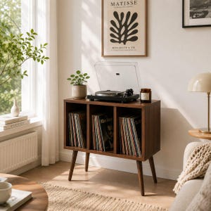 May include: A dark wood record player console with three open storage compartments filled with vinyl records. The console features four tapered legs and a black turntable with a clear dust cover placed on the top surface of the unit.