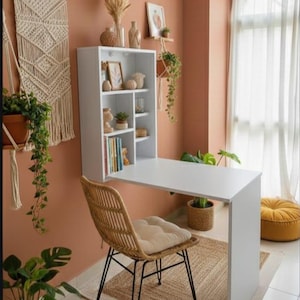 Puede incluir: Un escritorio blanco montado en la pared con una estantería a juego, una silla tejida y una alfombra de yute. La habitación está decorada con plantas, tapices de macramé y obras de arte enmarcadas. Las paredes están pintadas en un cálido color melocotón.