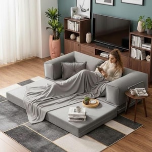 Puede incluir: Un sof&aacute; modular gris con una manta y un coj&iacute;n a juego. El sof&aacute; est&aacute; en un sal&oacute;n con una alfombra, muebles de madera y un televisor. Una persona se relaja en el sof&aacute;.