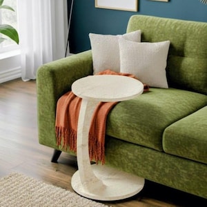 May include: A living room setting with a green sofa, two white textured cushions, and an orange fringed throw. A cream-coloured side table with a round top and curved base is positioned in front of the sofa. The room has wooden flooring and a neutral rug.