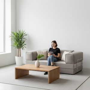 Puede incluir: Un sal&oacute;n moderno con un sof&aacute; esquinero gris claro, una mesa de centro rectangular de madera y una planta alta en maceta. Una persona est&aacute; sentada en el sof&aacute;, usando una tableta. La habitaci&oacute;n tiene un dise&ntilde;o minimalista con una paleta de colores neutros.