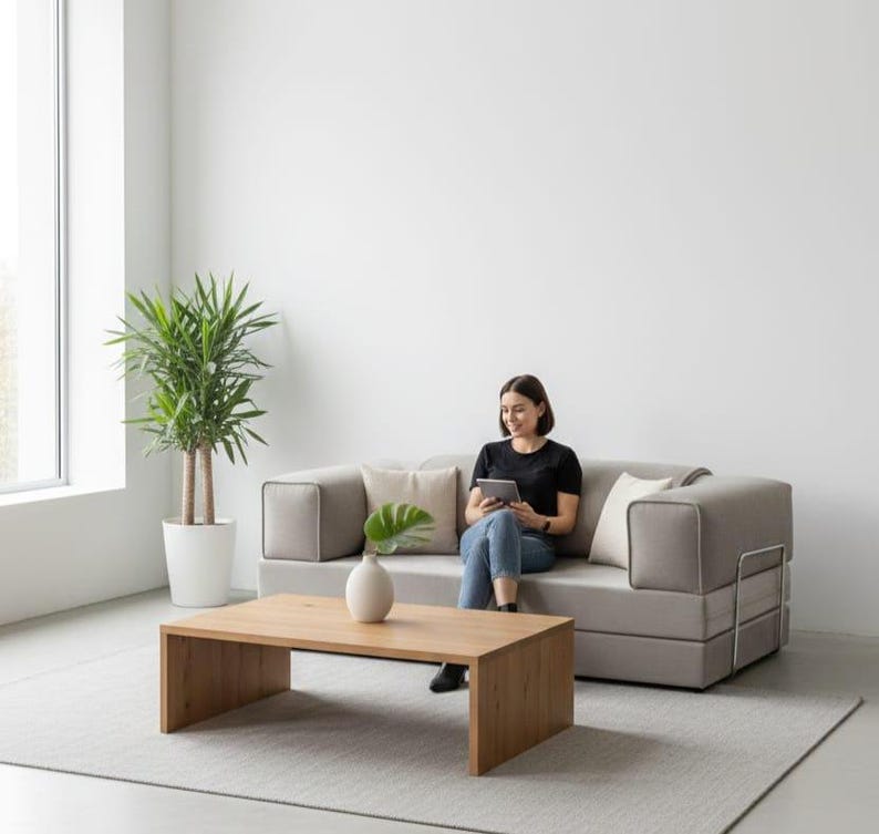 Puede incluir: Un sal&oacute;n moderno con un sof&aacute; esquinero gris claro, una mesa de centro rectangular de madera y una planta alta en maceta. Una persona est&aacute; sentada en el sof&aacute;, usando una tableta. La habitaci&oacute;n tiene un dise&ntilde;o minimalista con una paleta de colores neutros.