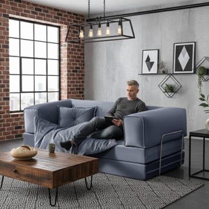 Puede incluir: Un sof&aacute; modular azul con una persona sentada en &eacute;l, una mesa de centro de madera y una mesa auxiliar negra. El sof&aacute; tiene una almohada y una manta. La mesa de centro tiene un cuenco decorativo y una taza. La habitaci&oacute;n tiene una pared de ladrillo y una ventana grande.