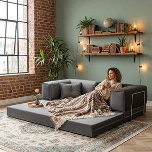 Puede incluir: Un sof&aacute; modular gris con una manta estampada y cojines. El sof&aacute; est&aacute; en un sal&oacute;n con una pared de ladrillo, una ventana grande y estanter&iacute;as de madera con libros y plantas.