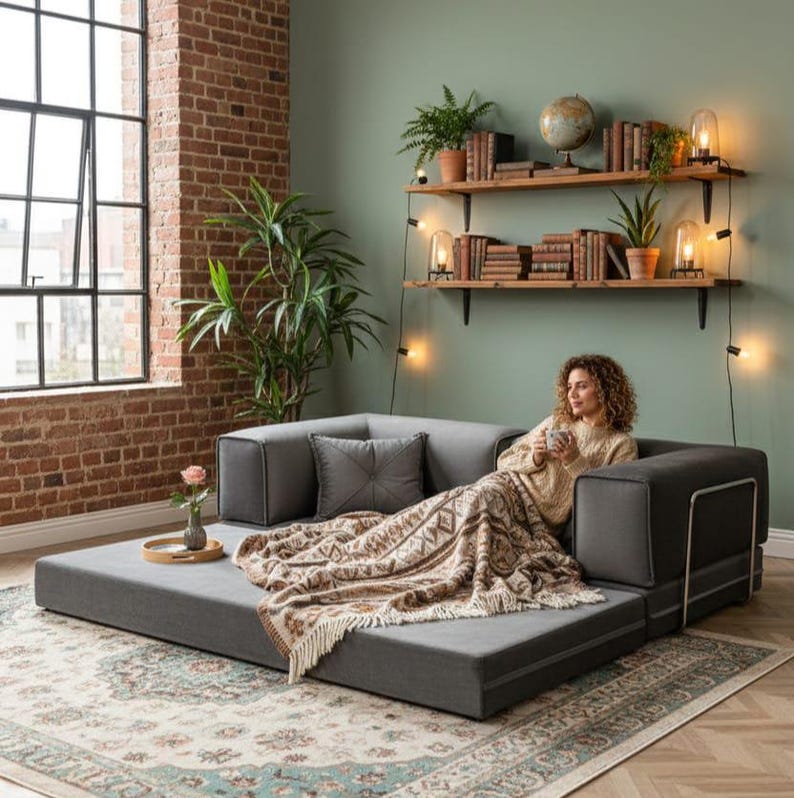 Puede incluir: Un sof&aacute; modular gris con una manta estampada y cojines. El sof&aacute; est&aacute; en un sal&oacute;n con una pared de ladrillo, una ventana grande y estanter&iacute;as de madera con libros y plantas.