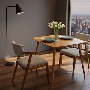 May include: A small, square wooden dining table with two chairs. The chairs have light-colored patterned upholstery and wooden frames. The table is set with plates and silverware. A vase of white tulips sits on the table. A black floor lamp is in the background.