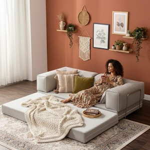 Puede incluir: Un sof&aacute; modular gris claro con una manta de punto color crema. Cojines decorativos en tonos neutros y una peque&ntilde;a bandeja de madera con un libro est&aacute;n en el sof&aacute;. El fondo presenta arte mural y plantas.