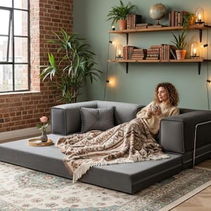 Puede incluir: Un sof&aacute; modular gris con una manta estampada y un coj&iacute;n decorativo. El sof&aacute; est&aacute; en un sal&oacute;n con una pared de ladrillo, una ventana grande y estanter&iacute;as de madera con libros y plantas.