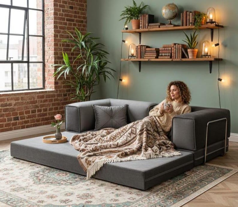 Puede incluir: Un sof&aacute; modular gris con una manta estampada y un coj&iacute;n decorativo. El sof&aacute; est&aacute; en un sal&oacute;n con una pared de ladrillo, una ventana grande y estanter&iacute;as de madera con libros y plantas.