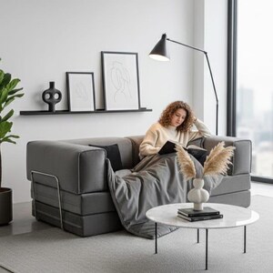 Puede incluir: Un sof&aacute; gris moderno con una persona leyendo un libro, una l&aacute;mpara de pie de metal negro y una mesa de centro redonda de m&aacute;rmol. Art&iacute;culos decorativos: estante negro con arte y planta en maceta.