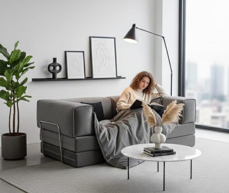 Puede incluir: Un sof&aacute; gris moderno con una persona leyendo un libro, una l&aacute;mpara de pie de metal negro y una mesa de centro redonda de m&aacute;rmol. Art&iacute;culos decorativos: estante negro con arte y planta en maceta.