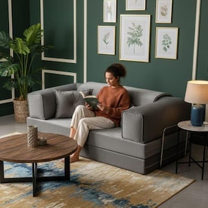 Puede incluir: Un sof&aacute; cama gris con una persona leyendo un libro. El sof&aacute; est&aacute; en una habitaci&oacute;n con una pared verde oscuro, estampados bot&aacute;nicos enmarcados y una mesa de centro redonda de madera. Una alfombra azul y dorada est&aacute; en el suelo.