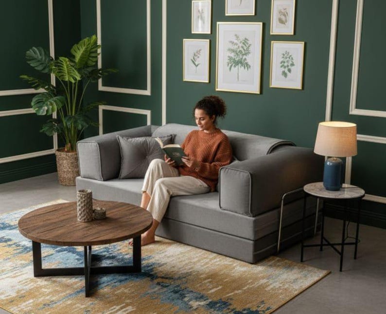 Puede incluir: Un sof&aacute; cama gris con una persona leyendo un libro. El sof&aacute; est&aacute; en una habitaci&oacute;n con una pared verde oscuro, estampados bot&aacute;nicos enmarcados y una mesa de centro redonda de madera. Una alfombra azul y dorada est&aacute; en el suelo.