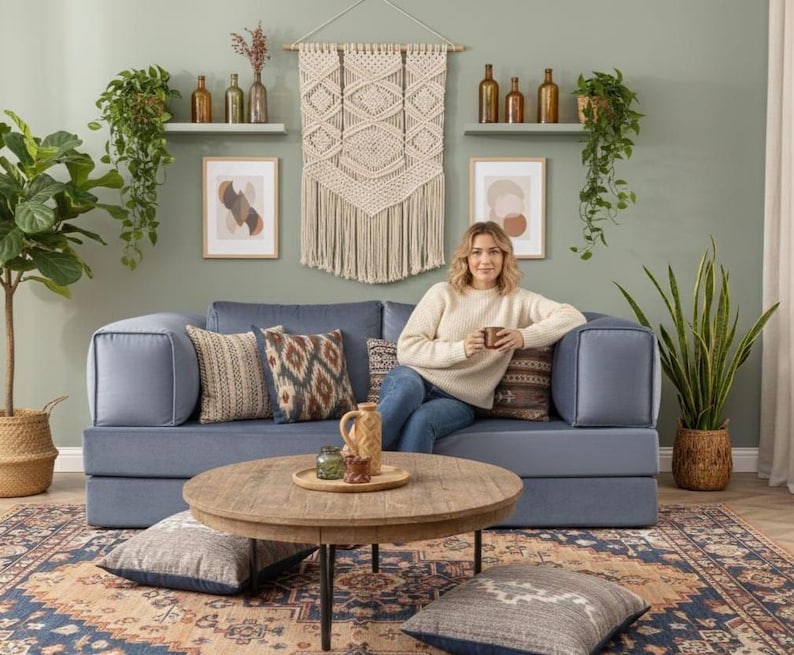 Puede incluir: Un sal&oacute;n acogedor con un sof&aacute; azul, cojines decorativos, una mesa de centro redonda de madera y una alfombra estampada. La habitaci&oacute;n est&aacute; decorada con plantas, obras de arte y un tapiz de macram&eacute;, creando un ambiente c&aacute;lido.
