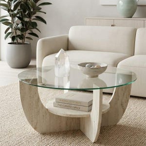 May include: A round coffee table with a clear glass top and a beige stone base. The base has a curved design with a lower shelf holding two books. A clear crystal and a decorative bowl sit on the table.