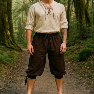 May include: A beige linen shirt with a laced neckline and brown drawstring shorts. The outfit is suitable for historical reenactments or themed events. The clothing is made of natural fabrics and is designed for comfort and style.