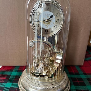 May include: A silver-toned Rhythm Quartz clock encased in a glass dome. The clock face is gold with black numerals and hands. The base is ornate, and a small carousel is visible inside. The clock sits on a red and green plaid surface.