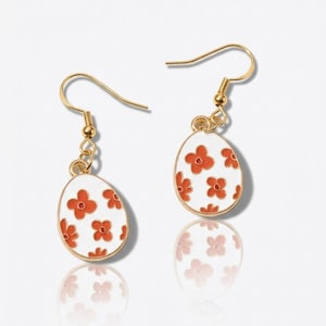 May include: A pair of gold-colored earrings featuring an egg-shaped pendant. The white pendant is decorated with orange flower designs and outlined in gold. The earrings have a hook closure.