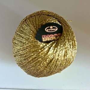 May include: A spherical ball of metallic gold yarn with a shimmering, reflective texture. A black circular label is centred on the top, featuring the white text STENLI and the word DISCO in large, bold, orange-toned letters with a dark outline.