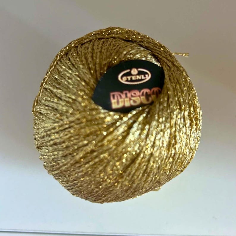 May include: A spherical ball of metallic gold yarn with a shimmering, reflective texture. A black circular label is centred on the top, featuring the white text STENLI and the word DISCO in large, bold, orange-toned letters with a dark outline.