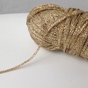 Può includere: Un gomitolo di filato metallizzato dorato con un filo che si estende verso l'esterno. Il filato ha un aspetto lucido e strutturato, che riflette la luce. Questo materiale artigianale è adatto per lavorare a maglia, all'uncinetto o altri progetti tessili.