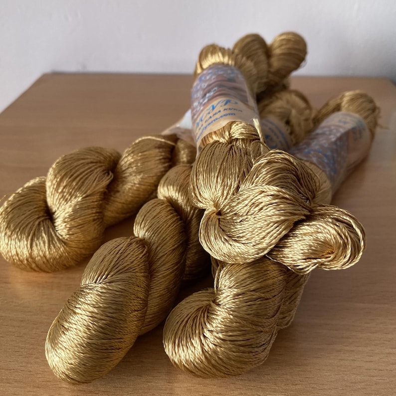 K&ouml;nnte beinhalten: Mehrere Str&auml;nge goldfarbenen Seidenfadens liegen auf einer Holzoberfl&auml;che. Der Faden ist eng zu B&uuml;ndeln gewickelt und hat eine gl&auml;nzende, glatte Textur. Einige Str&auml;nge sind mit Etiketten versehen.