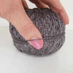 Può includere: Un gomitolo di filato grigio con fili argentati metallici. Una mano con smalto rosa tiene il filato, evidenziando la consistenza e i dettagli. Questo filato è adatto per progetti di maglieria o uncinetto.
