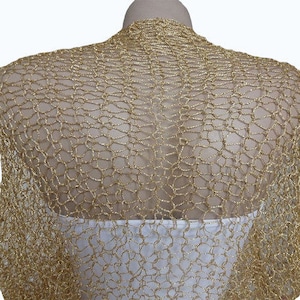 May include: A gold metallic bolero shrug featuring an open, loose-knit crochet pattern. The garment has a shimmering, textured finish and is draped over a white base layer, covering the shoulders and upper back with a lightweight, net-like structure.