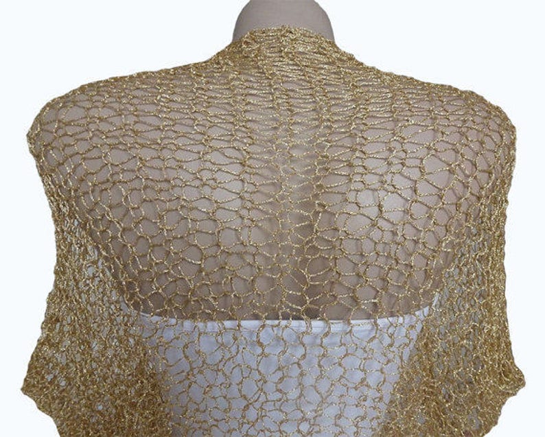 May include: A gold metallic bolero shrug featuring an open, loose-knit crochet pattern. The garment has a shimmering, textured finish and is draped over a white base layer, covering the shoulders and upper back with a lightweight, net-like structure.