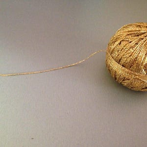 May include: A spherical ball of metallic gold yarn with a shimmering, reflective texture. A single strand of the gold thread extends outward from the main ball, showcasing the twisted, multi-ply construction of the metallic fibre material.