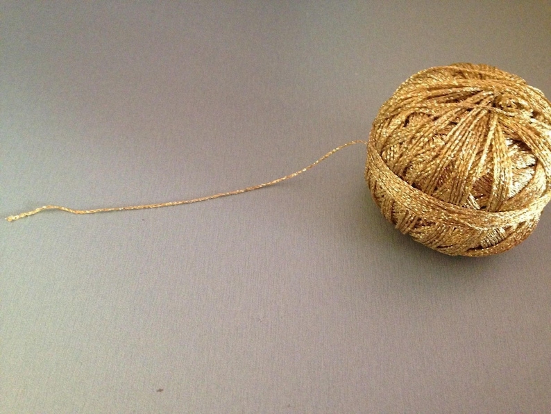May include: A spherical ball of metallic gold yarn with a shimmering, reflective texture. A single strand of the gold thread extends outward from the main ball, showcasing the twisted, multi-ply construction of the metallic fibre material.