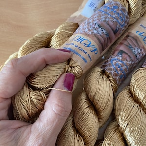 K&ouml;nnte beinhalten: Nahaufnahme mehrerer Str&auml;nge goldfarbenen Stickgarns. Das Garn ist eng gewickelt und hat einen leichten Glanz. Das Etikett auf dem Strang enth&auml;lt Text in einer Fremdsprache. Eine Hand mit bordeauxfarbenem Nagellack h&auml;lt einen Faden des Garns.